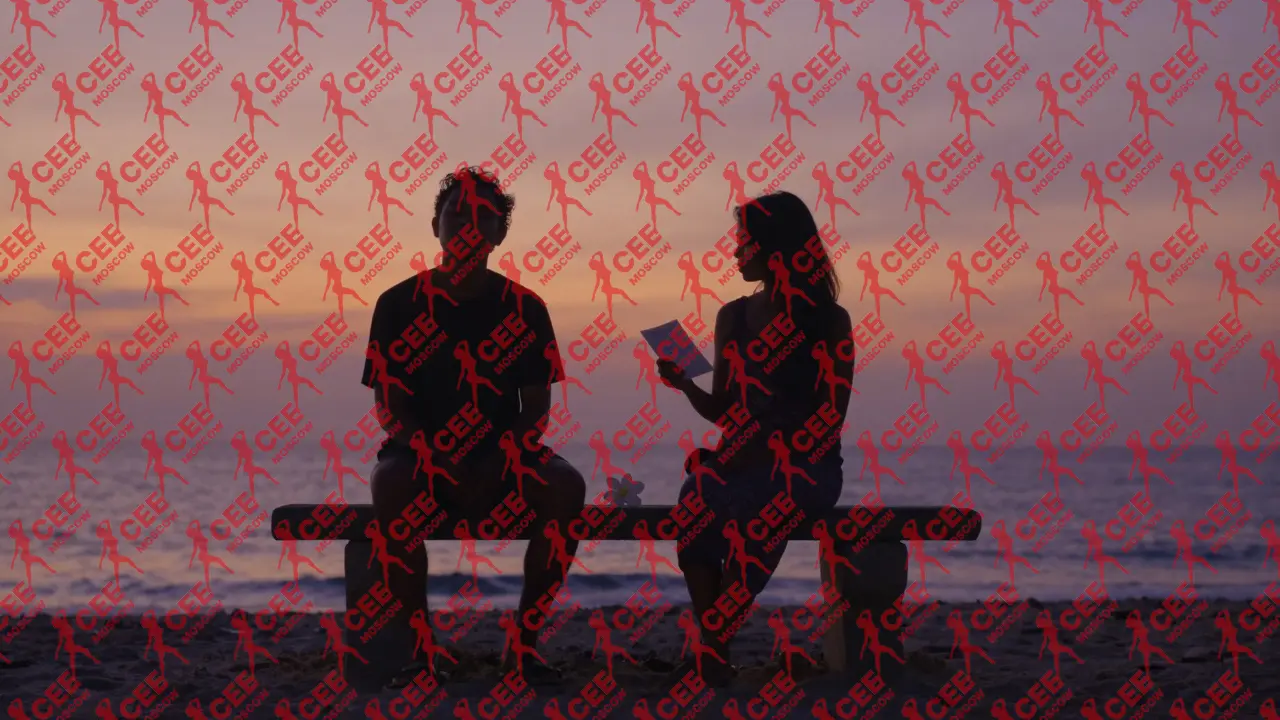 Двое сидят на берегу на закате, между ними лежит белый цветок, тишина и спокойствие.